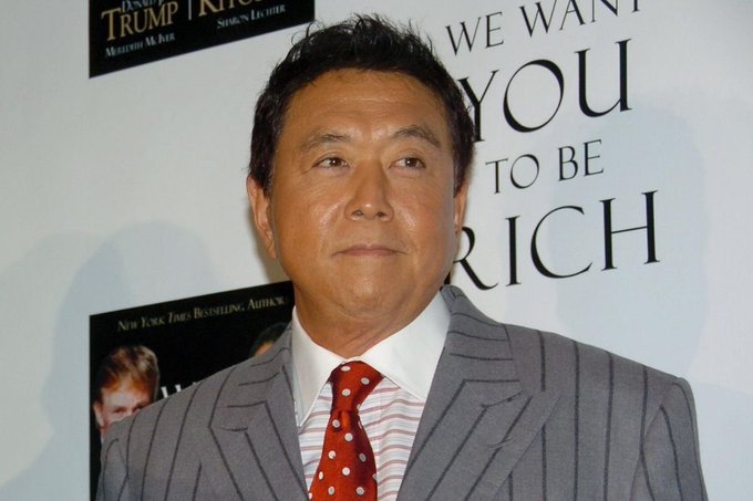 A man in a gray pinstripe suit with a red polka dot tie stands in front of a backdrop for the book 'We Want You to Be Rich' by Donald Trump and Robert Kiyosaki.