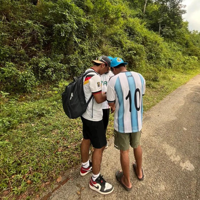 Two young men stand on a roadside wearing soccer jerseys—one in a Germany kit with a backpack and the other in an Argentina #10 'MESSI' jersey.