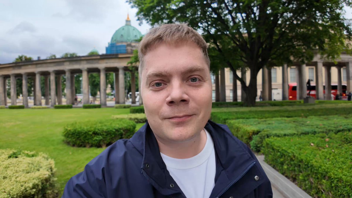 The image features a man wearing a blue jacket, standing next to a tree with a building in the background. The man appears to be posing for a photo or selfie, possibly at a popular tourist destination like Berlin. The image also contains text that reads "Mike booked a hotel in Berlin with @NOMADZxyz • saved ~~200$ compared to Expedia RAK rate - paid 5% less with USDC on @solana - love to bring real utility and see these stories". This suggests that the man is taking advantage of travel deals or discounts, possibly through a travel rewards program like USDC.