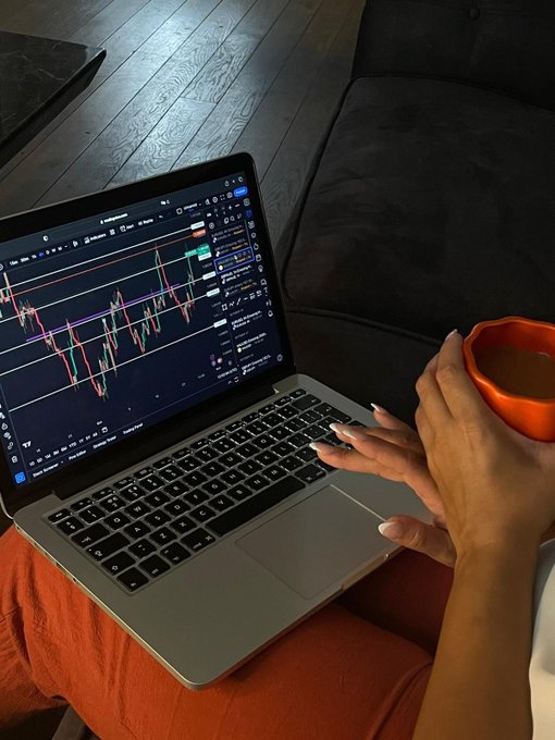 Person using a MacBook displaying a financial trading chart with candlestick patterns, while holding an orange mug, seated near a dark sofa.