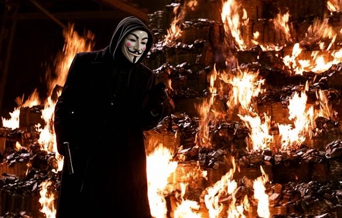 A figure wearing a black hooded coat and a Guy Fawkes mask stands in front of a large pile of burning debris and pallets at night.