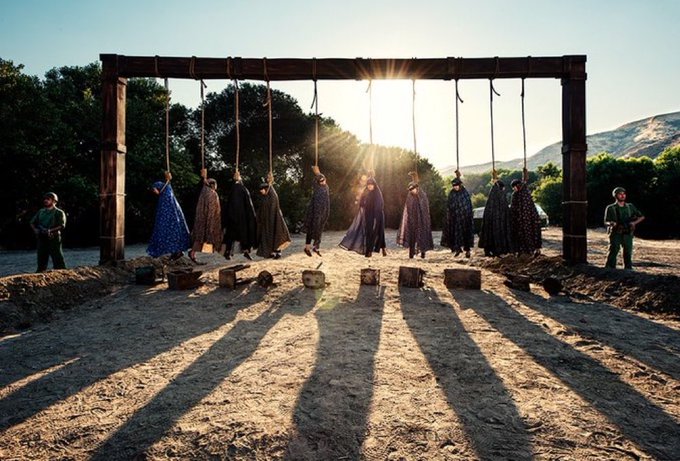 A dramatic scene of multiple people suspended by nooses from a wooden frame at sunset, with long shadows cast on the dirt ground.