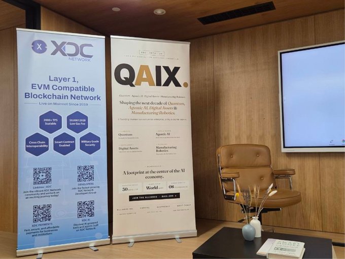 Two promotional banners stand in a wood-paneled room: one for XDC Network ('Layer 1, EVM Compatible Blockchain Network') and another for QAIX ('Shaping the next decade of Quantum, Agentic AI, Digital Assets & Manufacturing Robotics'), with a brown leather chair beside them.
