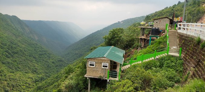 Scenic mountain landscape featuring small cabins with green roofs perched on a steep hillside overlooking a lush, misty valley.