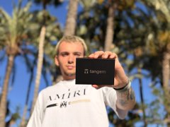 The image features a man holding up a black wallet with the word "Tangem" written on it, which suggests that he has chosen this particular wallet for its brand or design. The man appears to be posing for the camera while displaying his new purchase. The background of the photo shows palm trees and a clear blue sky, creating an outdoor setting. The image also contains text that reads: "people spend hours chasing 100x but 10 sec choosing a wallet priorities... self custody ... no seed phrases ... full control profit means nothing if you can't protect it". This message seems to be related to the man's decision to purchase the wallet and emphasizes the importance of making informed choices when it comes to personal belongings.
