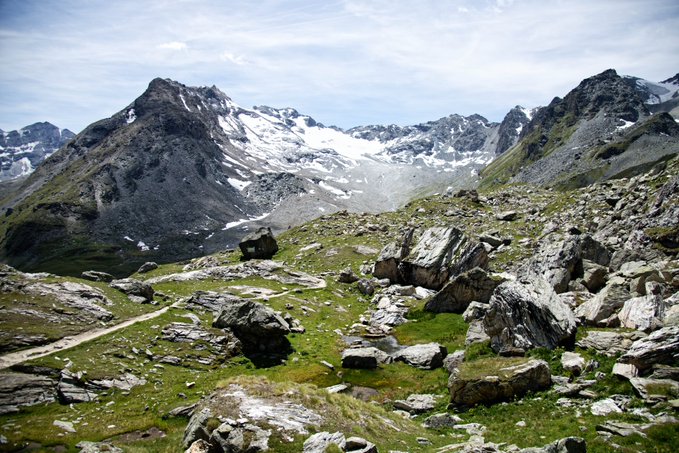 A scenic mountain landscape featuring rocky, snow-dusted peaks and green alpine meadows under a partly cloudy sky.