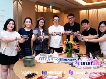 A group of eight people posing together at an event, holding items, with signage reading 'MEET OUR AI Web3', 'MEMECORE', and 'EVENT 3'.
