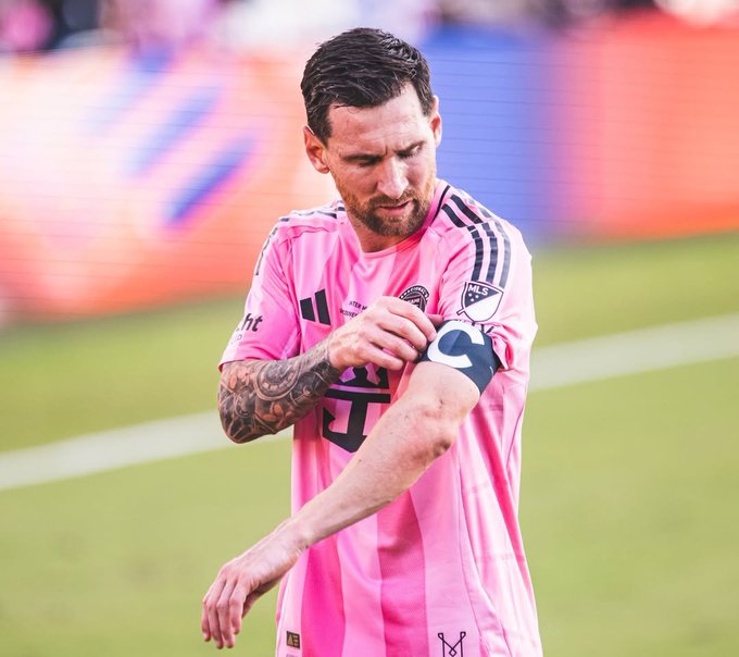 Soccer player Lionel Messi wearing a pink Inter Miami jersey, looking down while adjusting his captain's armband.