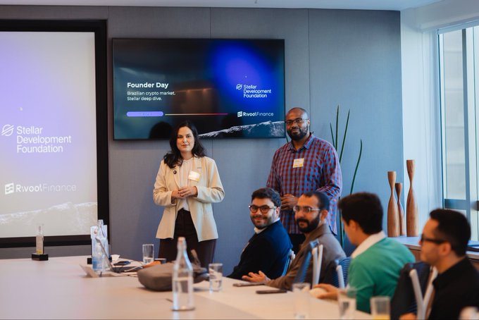 The image shows a group of people gathered around a conference table with a large screen displaying information about founder day at the Rovio Foundation. The attendees are engaged in conversation, possibly discussing the event or sharing their experiences related to blockchain technology. There is also a bottle on the table and a vase nearby, adding to the atmosphere of the meeting room.