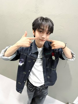 A young man with dark hair poses for a photo, making a playful gesture with both hands near his face while wearing a denim jacket over a white shirt. The image aligns with the "Take a V-sign pose photo" theme, as he strikes a casual, fan-like pose to celebrate the 1st July (7/1) date with ALPHADRIVEONE.