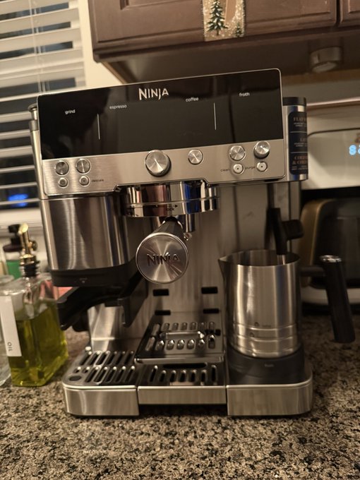 A Ninja brand stainless steel espresso machine sitting on a granite countertop, with a metal frothing pitcher attached to the side and window blinds visible in the background.