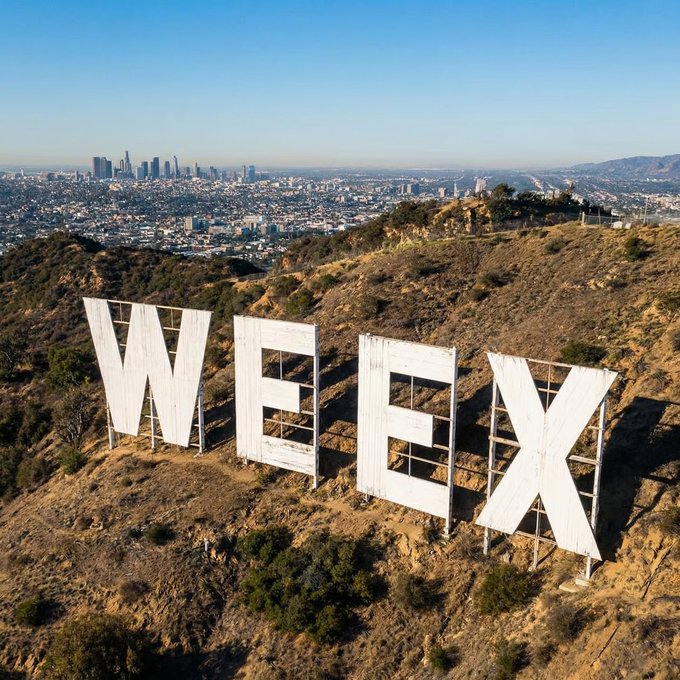 The iconic white "WEEEX" letters stand on a dry hillside with the LA skyline in the background, and the post's playful "GM!" (good morning) is fitting since the structure resembles the Hollywood Sign and greets viewers just like its world-famous counterpart greets Los Angeles every day.