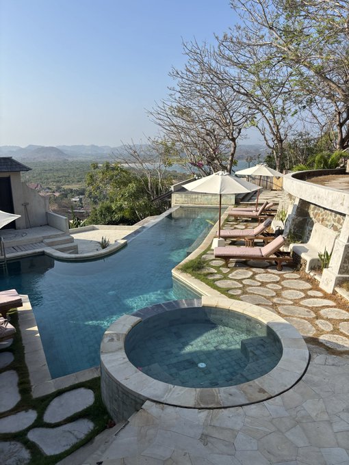 A scenic luxury resort features an infinity pool and a circular hot tub built from natural stone, with lounge chairs and umbrellas overlooking a vast mountainous landscape.