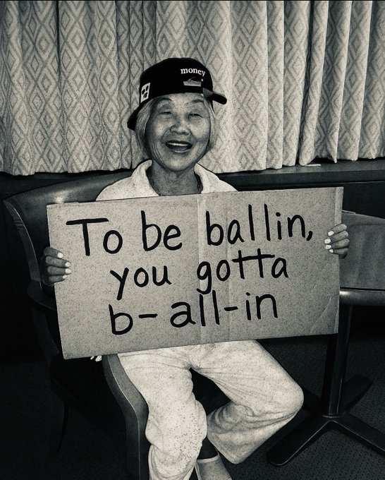 The image features a woman sitting down with her legs crossed while holding up a sign that says "To be ballin' you gotta b-all-in". The photo has been edited, giving it a vintage appearance. The caption above the picture reads "One thing I like about $RIVER is that there's always something happening. Whether it's new updates, community activity or fresh ideas from @River4fun and @RiverdotInc, the ecosystem never stands still."