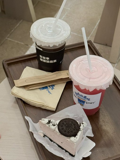 The image features a tray with two cups of coffee and a piece of cake sitting on top of it. One cup has a lid that appears to be made of plastic, while the other one has a lid made of paper. The cake also has a cookie on top of it. In the background, there is a chair visible, suggesting that this scene takes place in an indoor setting like a cafe or a restaurant.