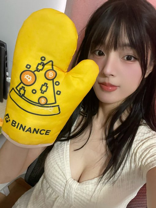 The image features a woman with long black hair wearing a white shirt, holding up a yellow oven mitt that has Bitcoin written on it. The woman appears to be posing for a photo or selfie, possibly sharing her enthusiasm for Bitcoin or promoting a product related to cryptocurrency.