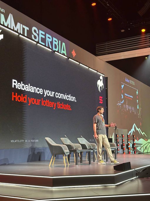 A speaker presents on a large stage at a 'SUMMIT SERBIA' event with a slide reading 'Rebalance your conviction. Hold your lottery tickets.'