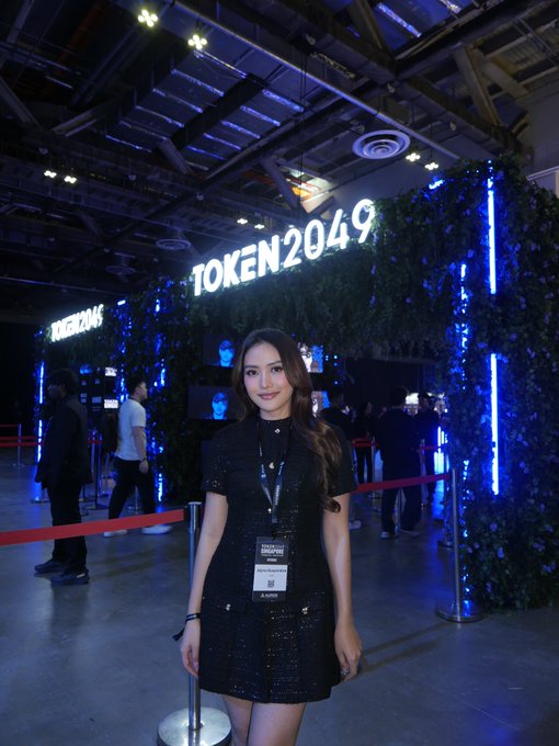 A woman in a black dress wearing a 'TOKEN2049' lanyard stands in front of the illuminated TOKEN2049 sign at what appears to be a conference venue.