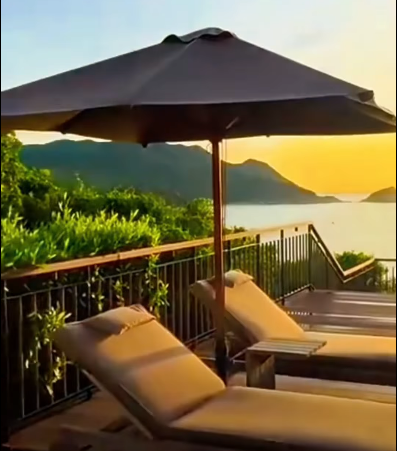 A serene balcony view at sunset featuring lounge chairs under a dark umbrella, overlooking lush green hills and a body of water.
