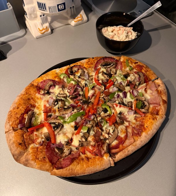 A large homemade pizza topped with salami, mushrooms, red onions, and bell peppers sits on a black plate next to a salad and R2-D2 salt and pepper shakers.