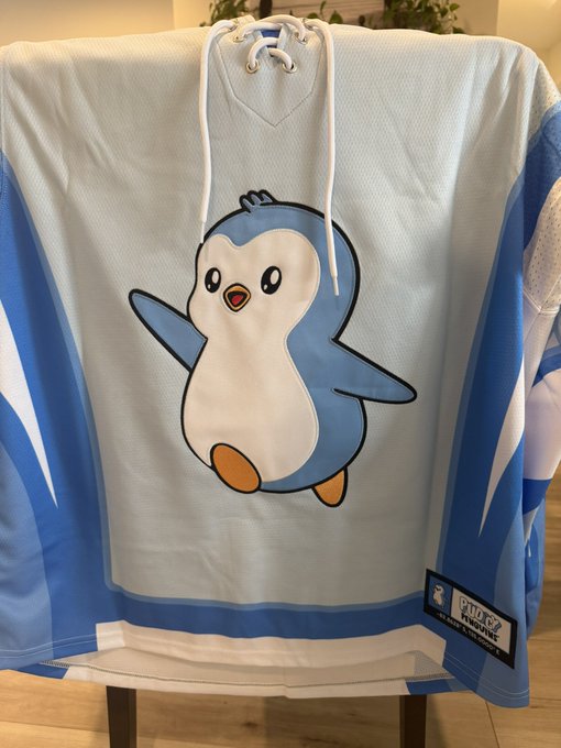 A white and blue hockey jersey displayed on a chair features a cute cartoon penguin (Piplup from Pokémon) graphic on the front, with a 'Pokémon' patch visible on the lower right hem.
