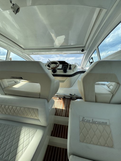 The interior of a white boat cabin features diamond-quilted leather seats with "Jean Turismo" embroidered on the backrest, a teak wood floor, and a sunlit dashboard.