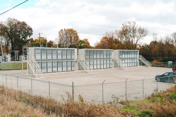Two-story stacked shipping container-style industrial buildings, likely mining infrastructure, sit behind a chain-link fence in a gravel lot with a black car parked nearby. The structures visually support the post's pitch about Bitcoin mining operations by depicting the kind of modular facility where hosted mining hardware could be housed and qualify for bonus depreciation.