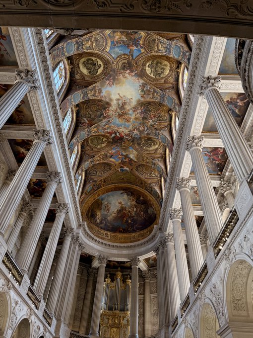 An upward view of an ornate palace or church interior showcasing Baroque architecture with tall columns, an elaborate gold-decorated ceiling featuring frescoes, and a golden pipe organ visible at the bottom.