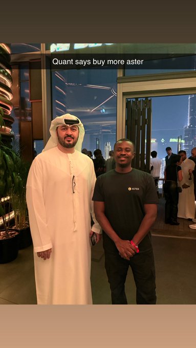Two men pose together indoors; one wears a traditional white thobe and ghutra while the other wears a dark ASTER t-shirt, with text overlay reading 'Quant says buy more aster'.