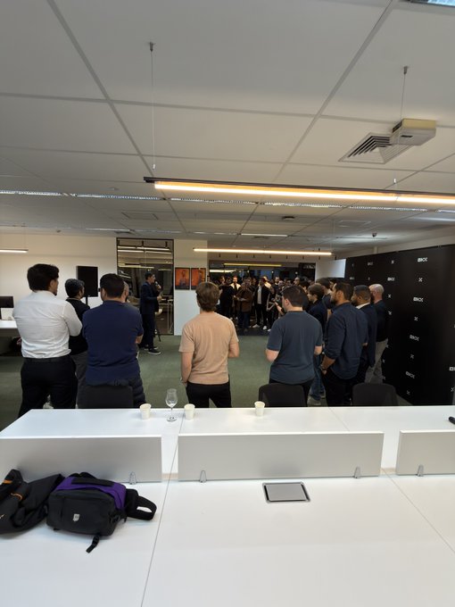 An office gathering with attendees standing in a conference room, featuring a camera and backpack on the front table.