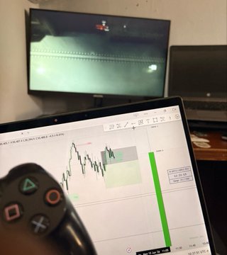 A person holds a PlayStation controller while a laptop displays a stock trading chart, with a TV in the background showing a video game.