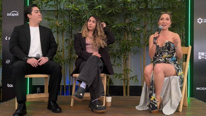 Three people sitting on a panel stage with microphones; a man in black on the left, a woman in a brown blazer in the middle, and a woman in a floral dress speaking on the right, with sponsors like 'bitspot' visible.