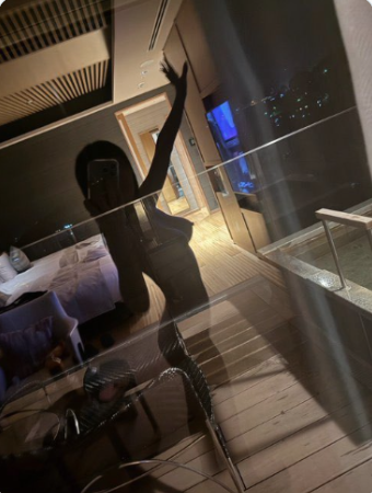 A low-light reflection photo of a person taking a mirror selfie on a wooden balcony, holding a phone with one arm raised, with bedroom visible in the background.