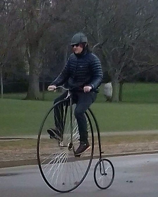 A man wearing a black jacket, sunglasses, and a cap rides a vintage penny-farthing bicycle on a paved path near a grassy park area.