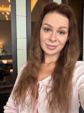 A woman with long brown hair wearing a light pink blouse takes a selfie in what appears to be a hotel or lounge setting with chandelier lighting in the background. The image conveys a professional yet approachable vibe, fitting her post about transitioning out of unemployment and seeking a role in Web3, AI, community, or content within the tech industry.