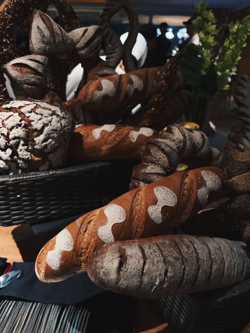 A close-up of an assortment of artisanal breads displayed in woven baskets, featuring rustic loaves with dark crusts, decorative scoring, and flour-dusted surfaces.
