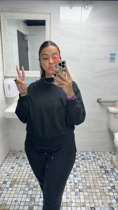 Mirror selfie of a woman in a black sweatshirt and black sweatpants standing in a tiled bathroom, holding a phone with stickers and making a peace sign, with a pink flower tucked behind her ear.