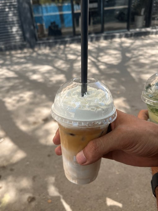 A hand holds a plastic cup of iced coffee topped with whipped cream and a black straw, with another green drink visible in the background on a sunny street.