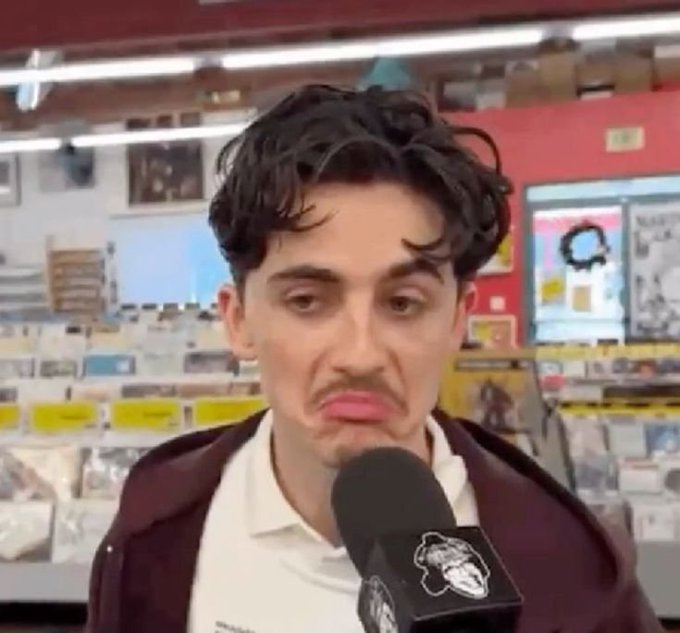 A young man with dark hair and a drawn-on mustache, looking displeased while being interviewed with a microphone in what appears to be a record store.