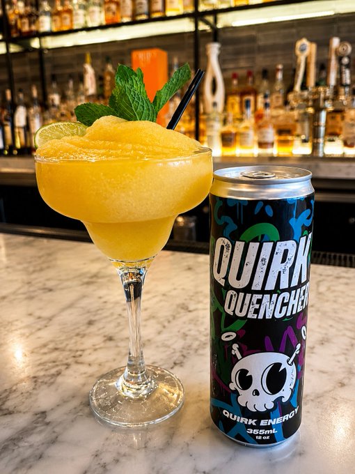 A vibrant orange frozen margarita garnished with fresh mint and lime sits on a marble bar top next to a 355ml can of "Quirk Quencher Quirk Energy" drink, featuring the brand's signature skull mascot in front of a well-stocked liquor shelf. The image showcases the new signature cocktail created using Quirk Energy, as highlighted in the post about the collaboration between Elephant and Dove restaurant and the Quirk energy drink brand.