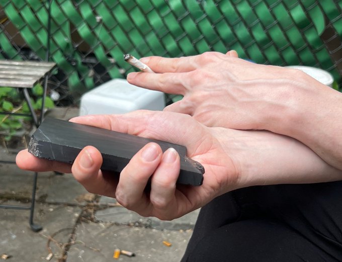 A pair of hands carefully holds a broken black smartphone and a lit cigarette, capturing a moment of reluctant disconnection as the person tries to step away from their device for a smoke break. The image humorously illustrates the post's critique of phone dependency, showing the absurdity of checking a cracked phone while trying to take a moment away from it.