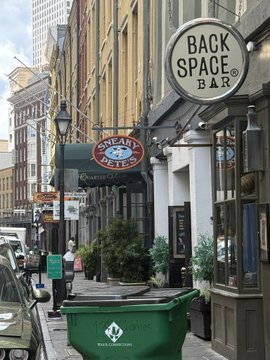 A street view showing a green dumpster in the foreground, with signs for 'BACK SPACE BAR' and a 'ONE WAY' sign mounted on buildings in what appears to be a downtown area.