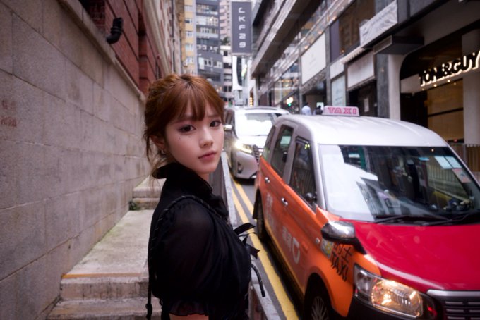 A young woman with brown hair and bangs wearing a black sheer top stands on a city street next to a red and orange taxi.