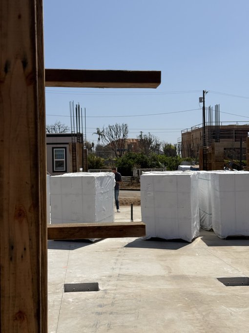 A view from a construction site looking out between wooden framing, showing a person standing among stacks of white-wrapped building materials on a concrete slab, with other buildings under construction in the background.