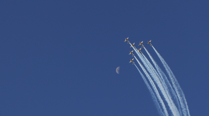 A formation of small airplanes flies through a deep blue sky, leaving white smoke trails behind them, with a pale crescent moon visible in the background.