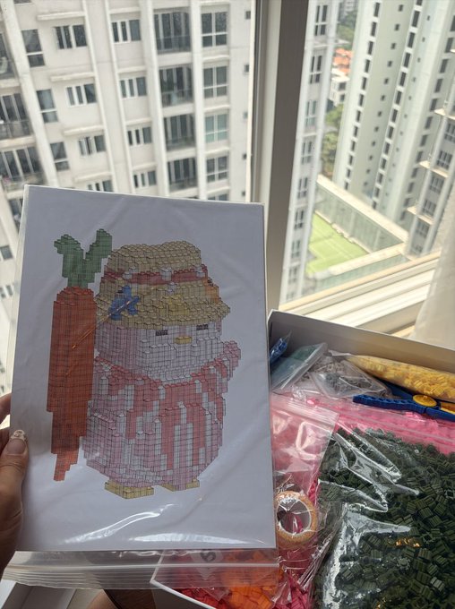 A person holding a printed pixel-art template of a character wearing a hat and cape, with bags of colored beads/blocks visible beside it, set against a window overlooking apartment buildings.