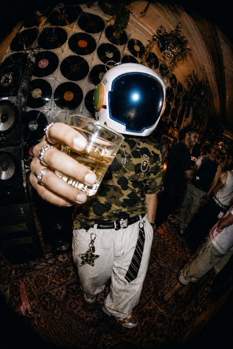 A person wearing an astronaut helmet and camouflage t-shirt holds out a glass of whiskey at a party venue decorated with vinyl records on the walls and speakers.