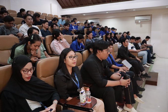An audience of young people seated in a tiered lecture hall or auditorium, with many using phones and laptops, while a woman in the foreground wears a black hijab and glasses.
