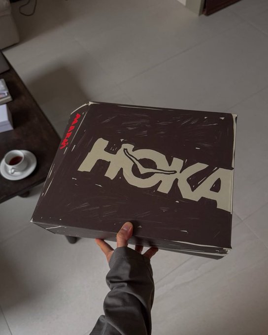 A person holds up a black HOKA shoe box, displaying the prominent gold "HOKA" logo on top, with what appears to be a coffee cup on a table visible in the background.