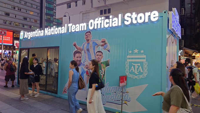 The image shows a group of people walking past an official store for Argentina National Team, which has a blue sign with white text advertising their merchandise. The store appears to be located in a shopping plaza or street market area, as there are several people gathered around it. One person is carrying a handbag and another is wearing a backpack.